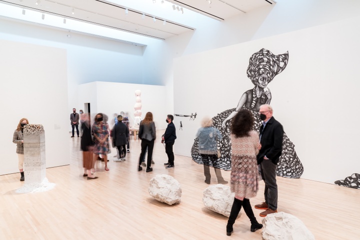 Wide view of gallery space with people moving through it. Visible are an installation of grey boulders on the floor and a large-scale black and white drawing of a woman in a huge skirted dress and a head turban, covering a half-wall inside the gallery.