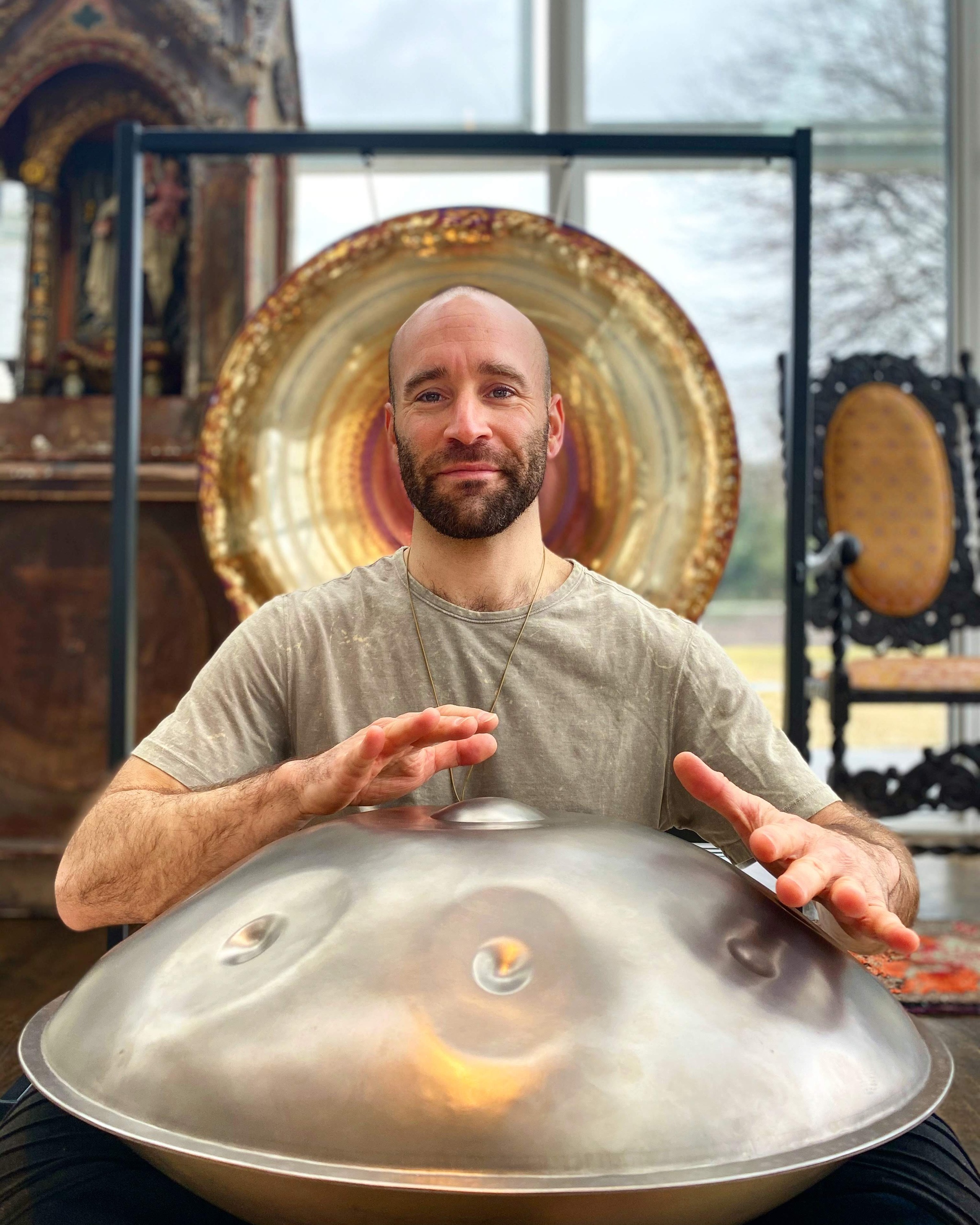 A white man smiles from behind a large metal percussion instrument that looks like a wok turned upside down. His hands are suspended over it as he drums on it. He wears a gray tshirt, smiles at us, and has a beard with a clean shaven head. Behind his head hangs a gong that surrounds his head like a halo. 