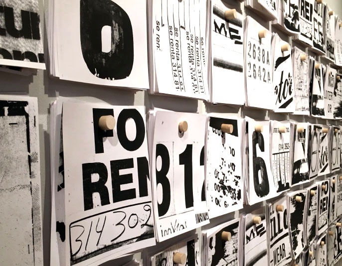 A close-up of a wall covered by sheets of paper hanging on wooden pegs. The papers, risograph prints, are white with black ink and depict parts of street signage.