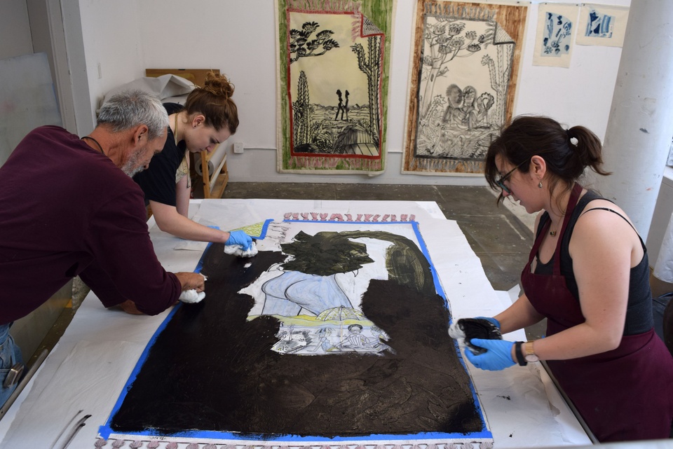 3 people rubbing black ink onto a large print on the table 