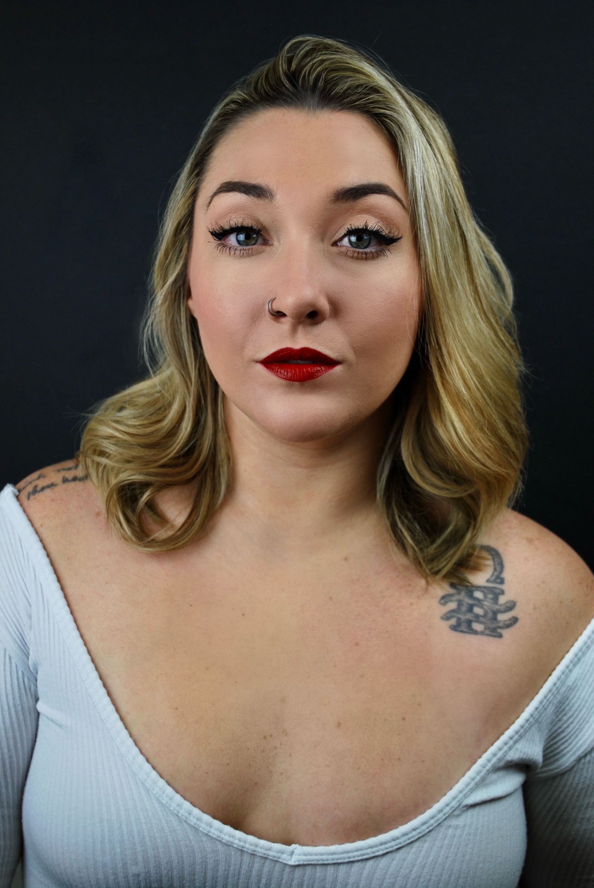 A portrait of the singer STONES, who has wavy blond hair that falls to her shoulder. She raises an eyebrow archly, and wears a low cut blue shirt that shows tattoos on both her shoulders.