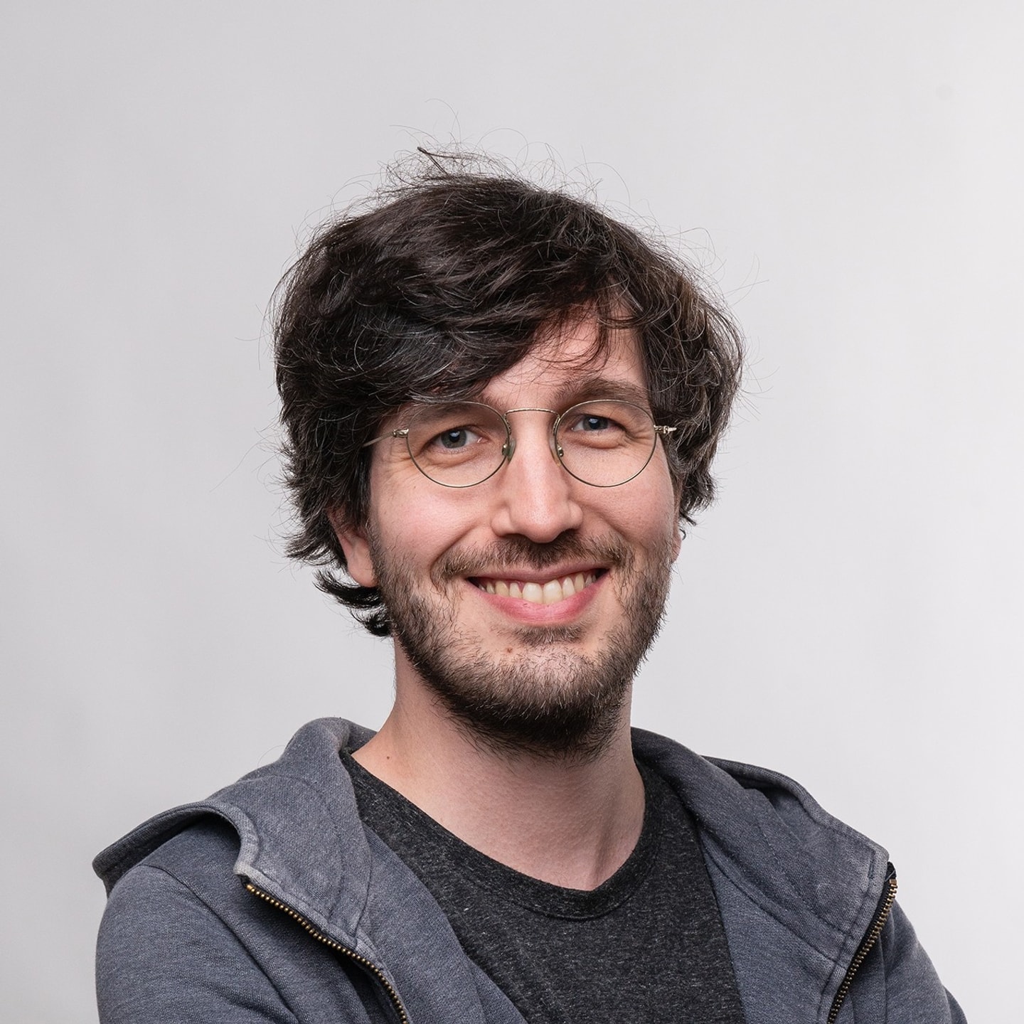 headshot of man with dark shaggy hair, short beard and round glasses wearing a black shirt and gray jacket against a light background