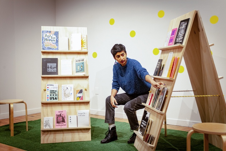 Shreyas sit between two magazine stands with various publications on them 