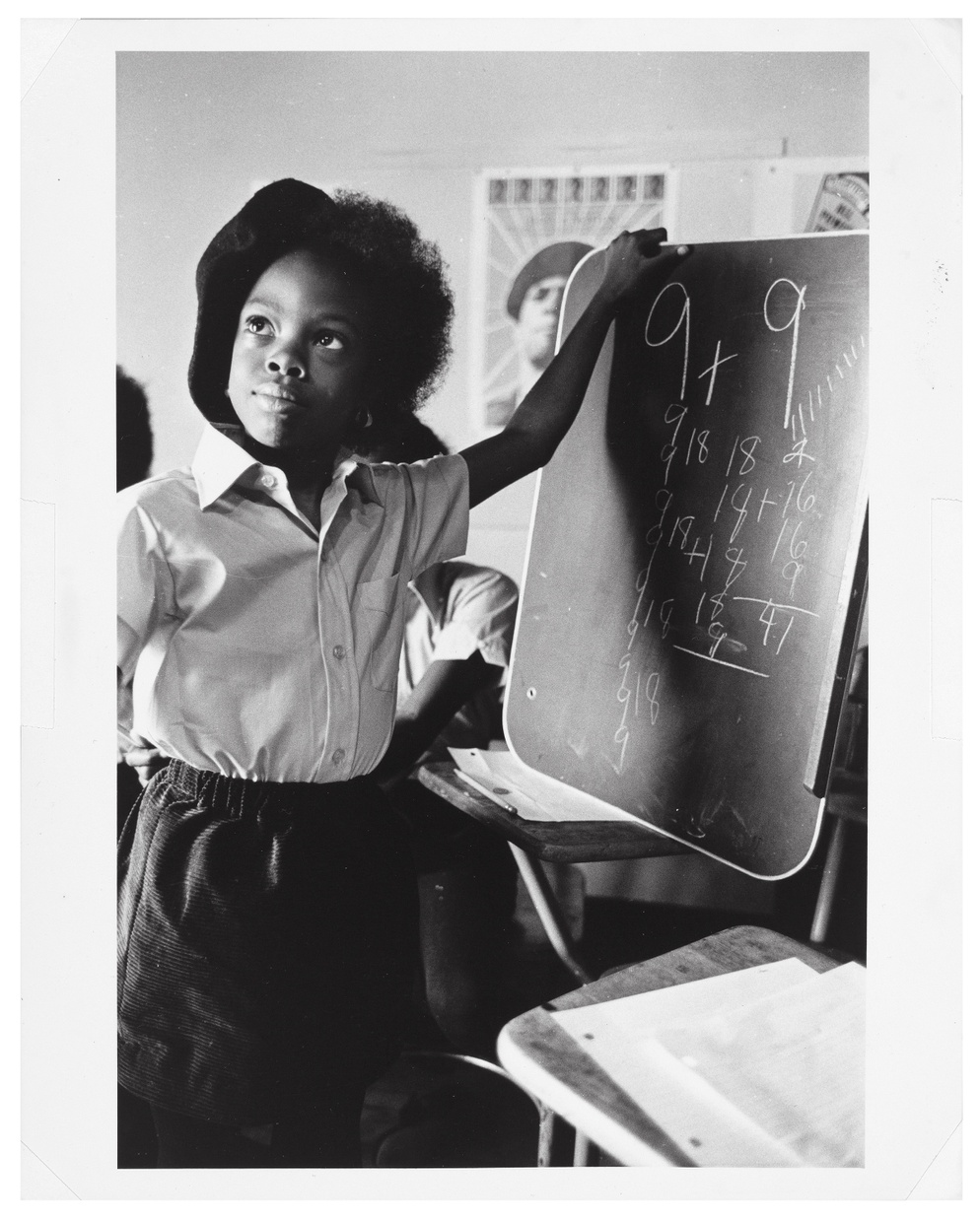 A black and white photograph of a little, Black girl wearing a school uniform and a beret holding up a small chalkboard with math equations written on it.