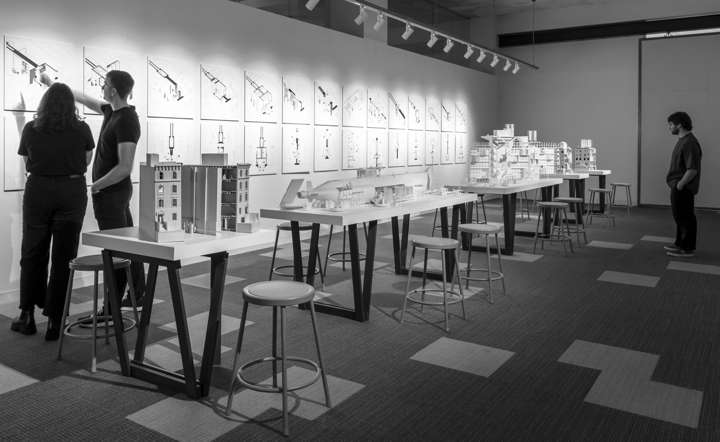 Black and white photo of an architecture exhibition installation. Two people in the foreground are pointing at a drawing on on the wall. Another in the background is looking at an architectural model.