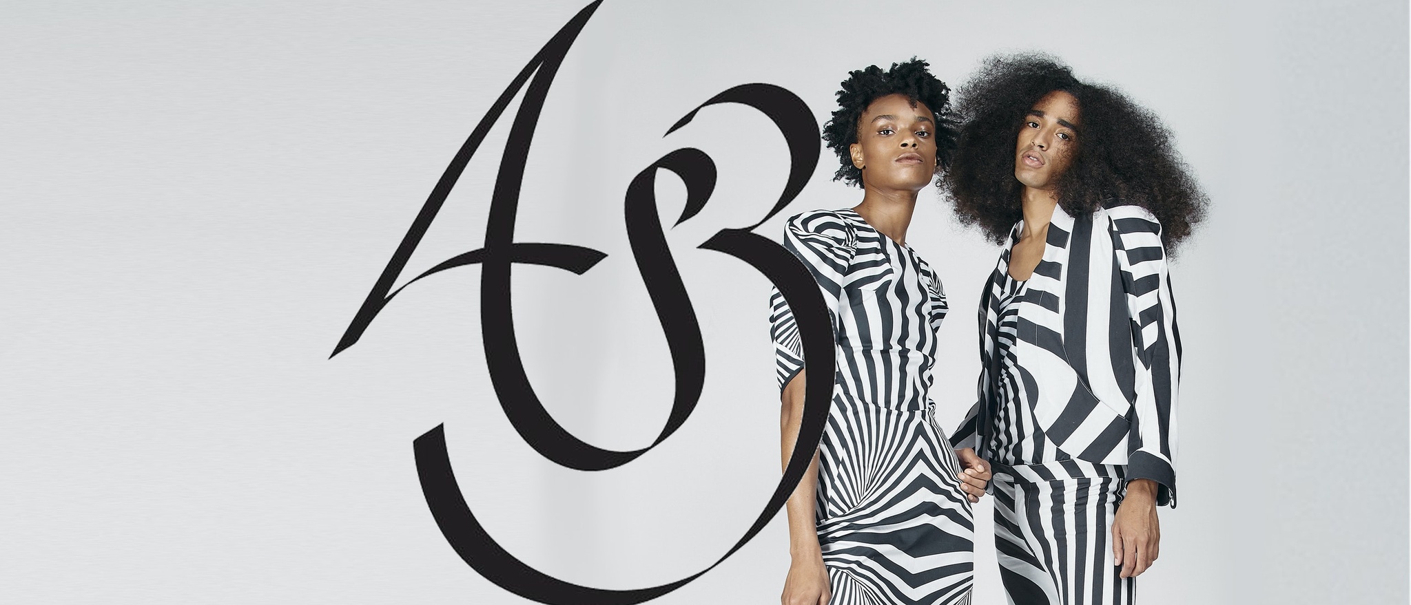 Two Black people dressed in outfits with bold black and white patterns that recall optical illusion patterns stand alongside a stylized script that looks like the cursive letters A and B