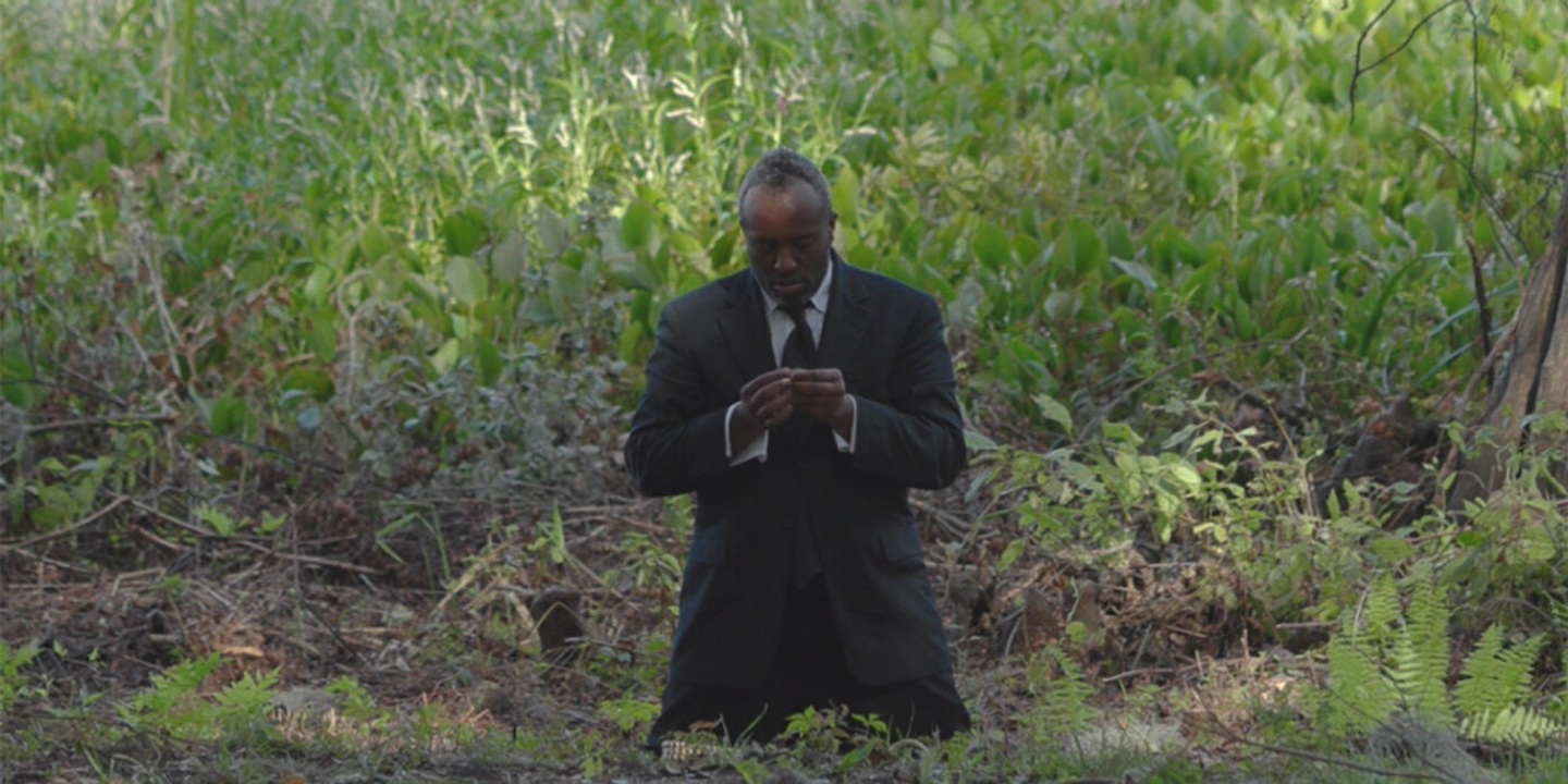 A man dressed in a full suit is kneeling in an outdoor area thick with brush, sticks, and green plants. The person's head is bowed somewhat, eyes closed, hand cupped with fingertips touching one another.