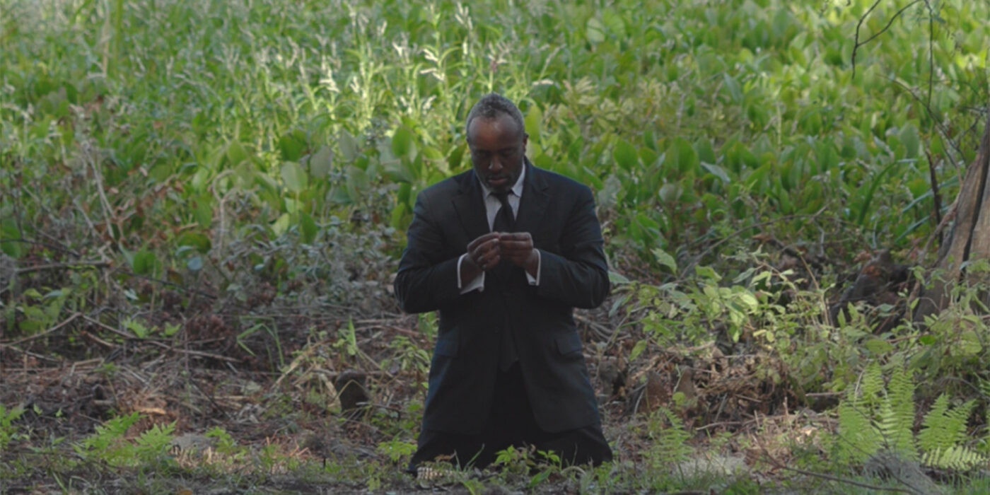 A man dressed in a full suit is kneeling in an outdoor area thick with brush, sticks, and green plants. The person's head is bowed somewhat, eyes closed, hand cupped with fingertips touching one another.