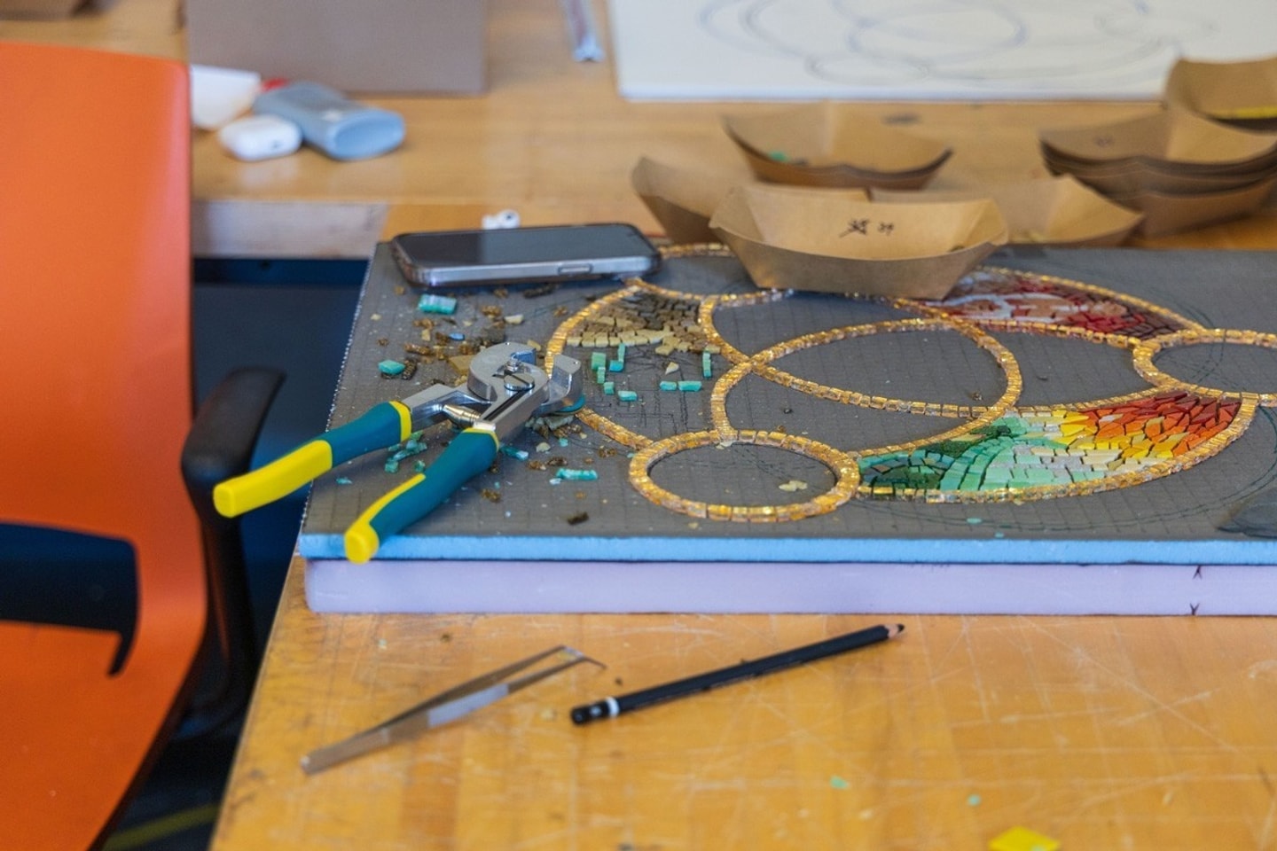 A mosaic artwork in progress on a wooden workbench, surrounded by mosaic tile nippers, tweezers, paper trays with tiles, and a pencil.