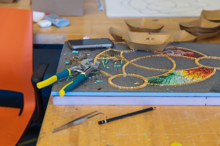 A mosaic artwork in progress on a wooden workbench, surrounded by mosaic tile nippers, tweezers, paper trays with tiles, and a pencil.