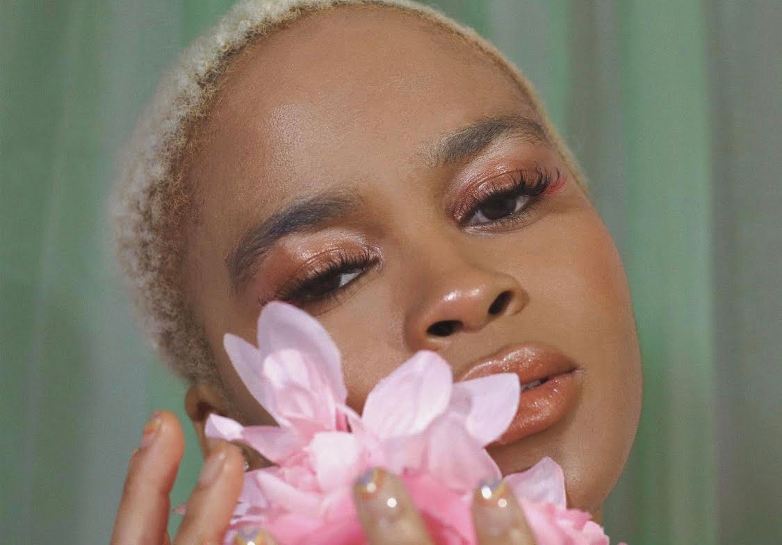 A Black woman with short blond hair seen in a close-up holding a pink flower in front of the lower half of her face. 