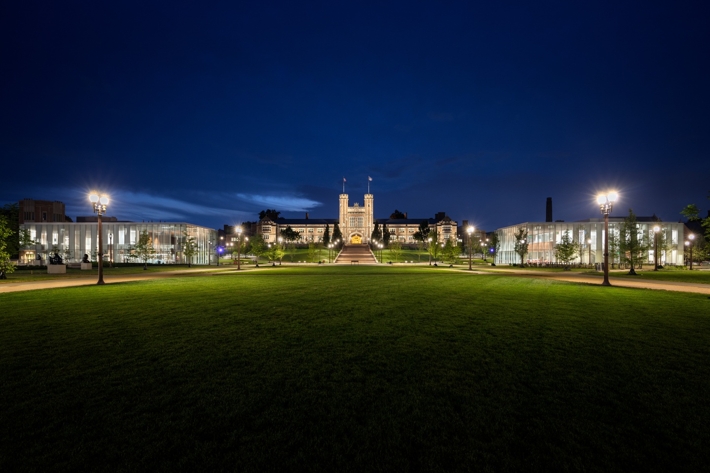 Nightime view of an expansive green space for WashU's new Tisch Park. Two glazed pavilions are lit up in the distance, flanking a collegiate-gothic building in the center.