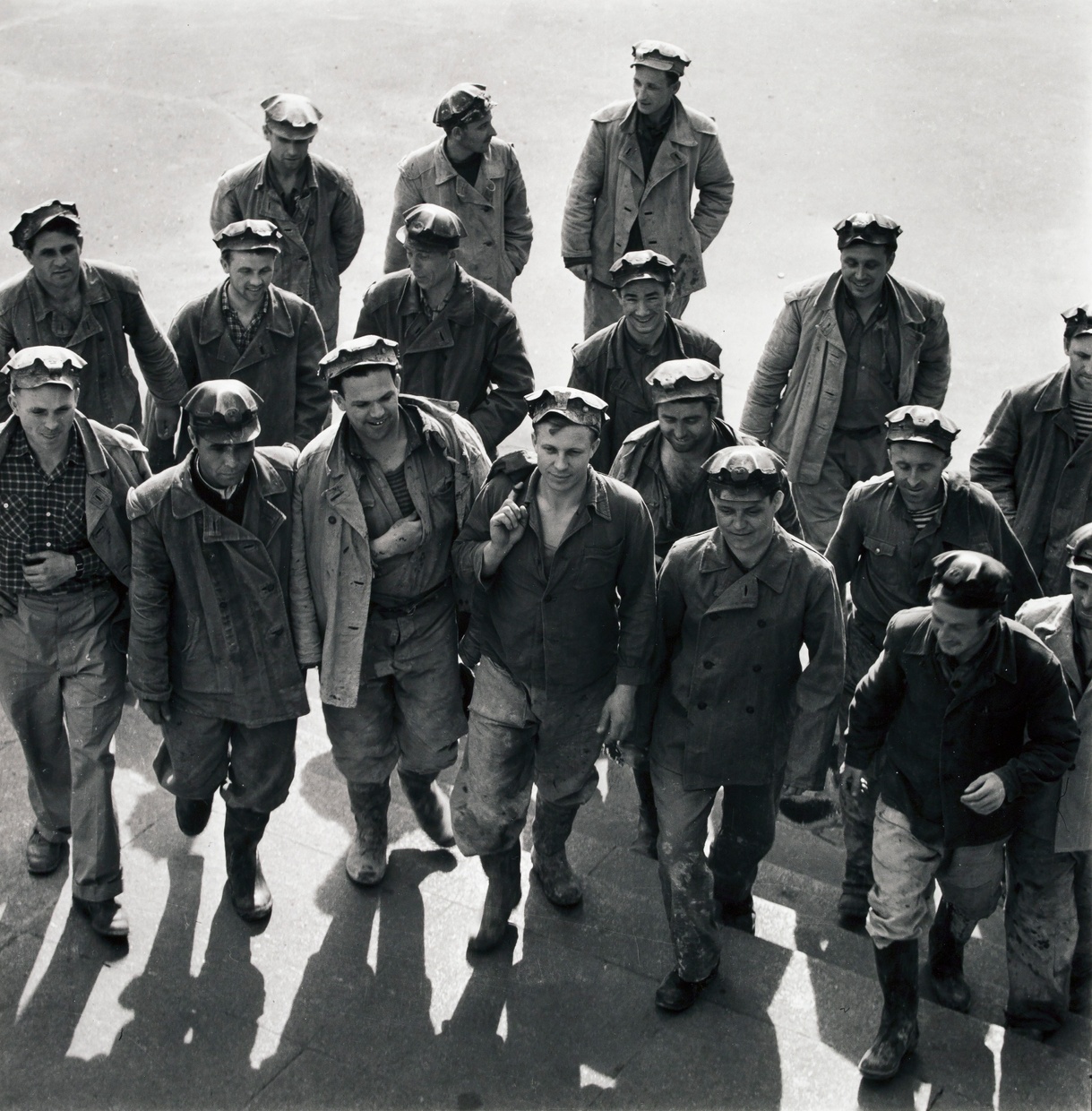 Black and white photograph of a group of 18 men walking up steps, wearing hats and dirty work clothes and boots.