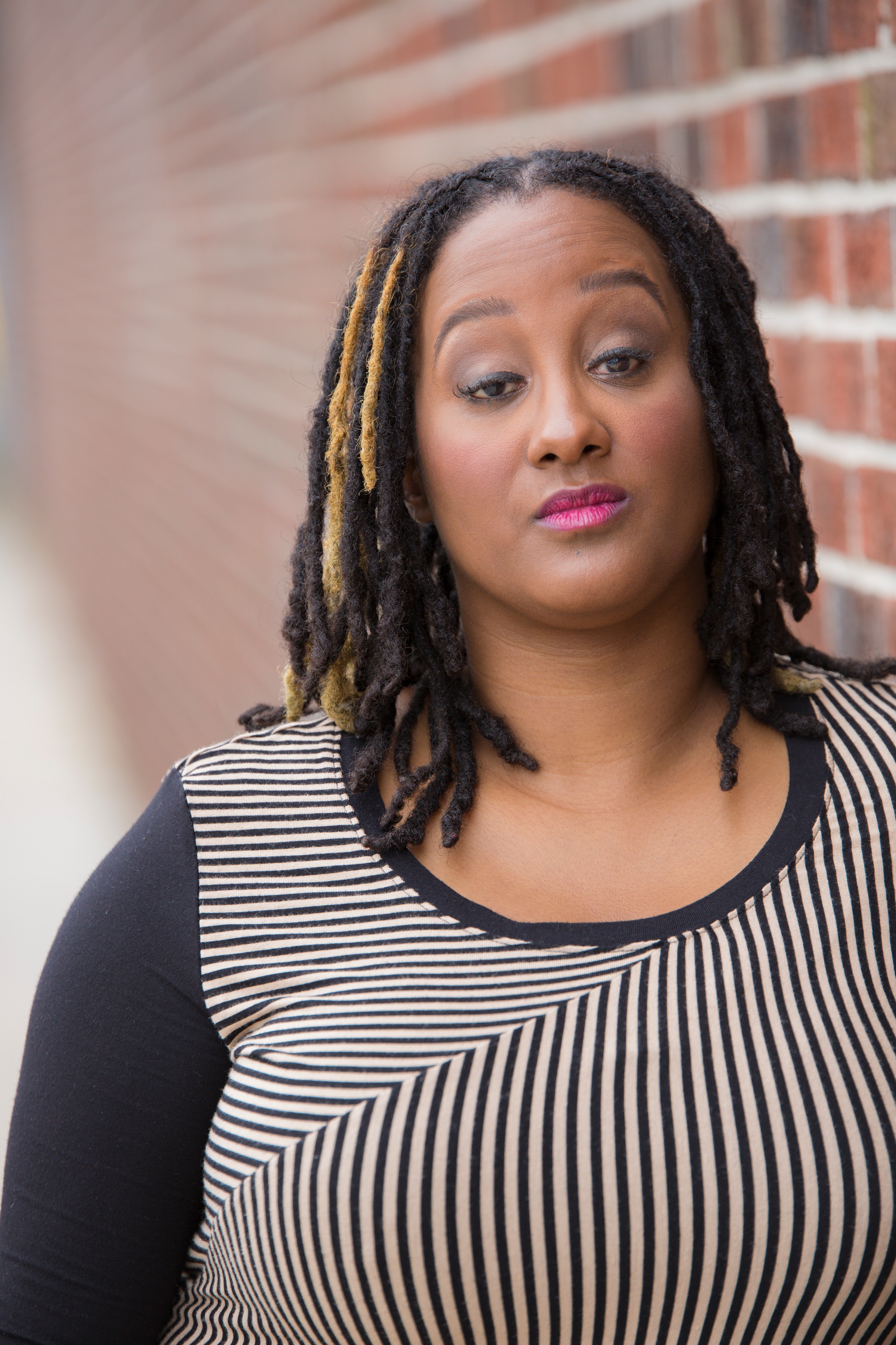 This is a headshot of Kayla Hamilton, who is a dark brown-skinned Black woman. She is posing in front of a blurred brick wall. She is wearing a long sleeve black & white striped shirt. She has light makeup and her gaze is towards us. Her black & golden highlighted dreads are down.] Photo by: Travis Magee