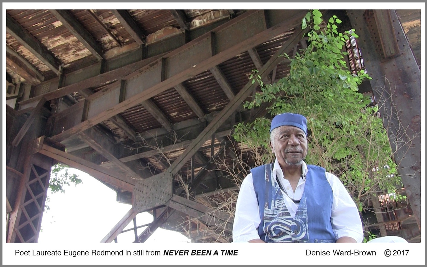Text reads, "Poet Laureate Eugene Redmond in still from NEVER BEEN A TIME" above a still from the video, in which Redmond wears a white shirt with ea blue vest and cap, under the floor of a wooden structure. There is a tree behind him.