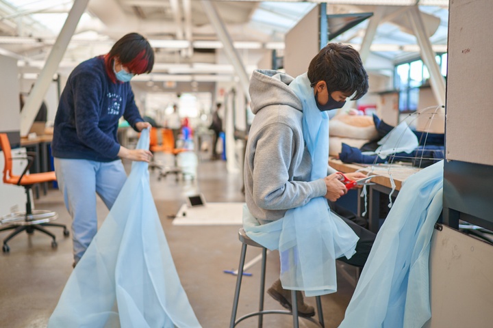Two students working in a light-filled studio space, cutting and adjusted a long, pale blue piece of fabric.