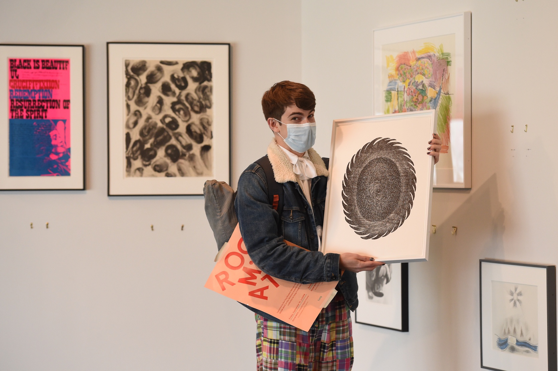 A masked student holds a framed artwork with a poster under their arm, and artwork hung on the walls behind them.