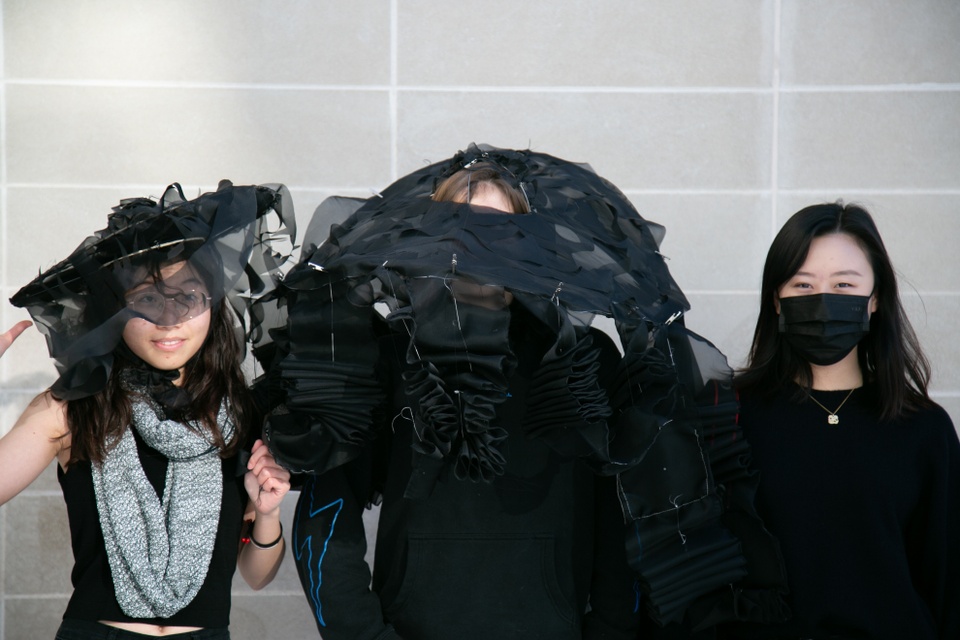 Three people standing against a gray wall, two of whom are wearing a shared black hat with lots of draping.