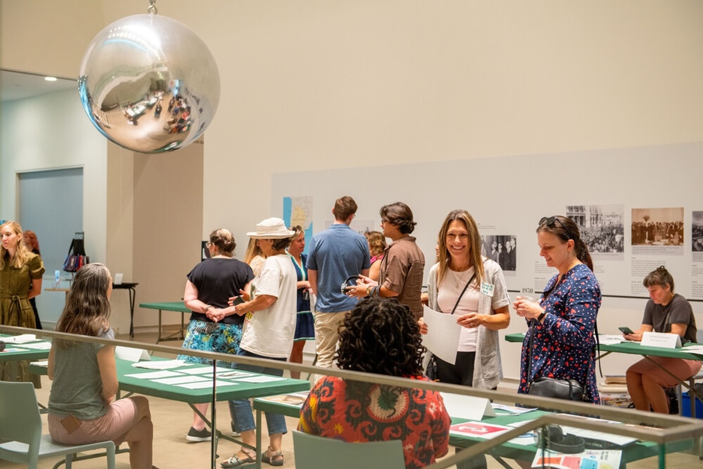 A group of visitors gathers in the atrium of the Kemper Art Museum, with tables displaying printed materials in the foreground and a historical timeline on the wall behind them. A large mirrored sphere hangs from the ceiling above the gathering.