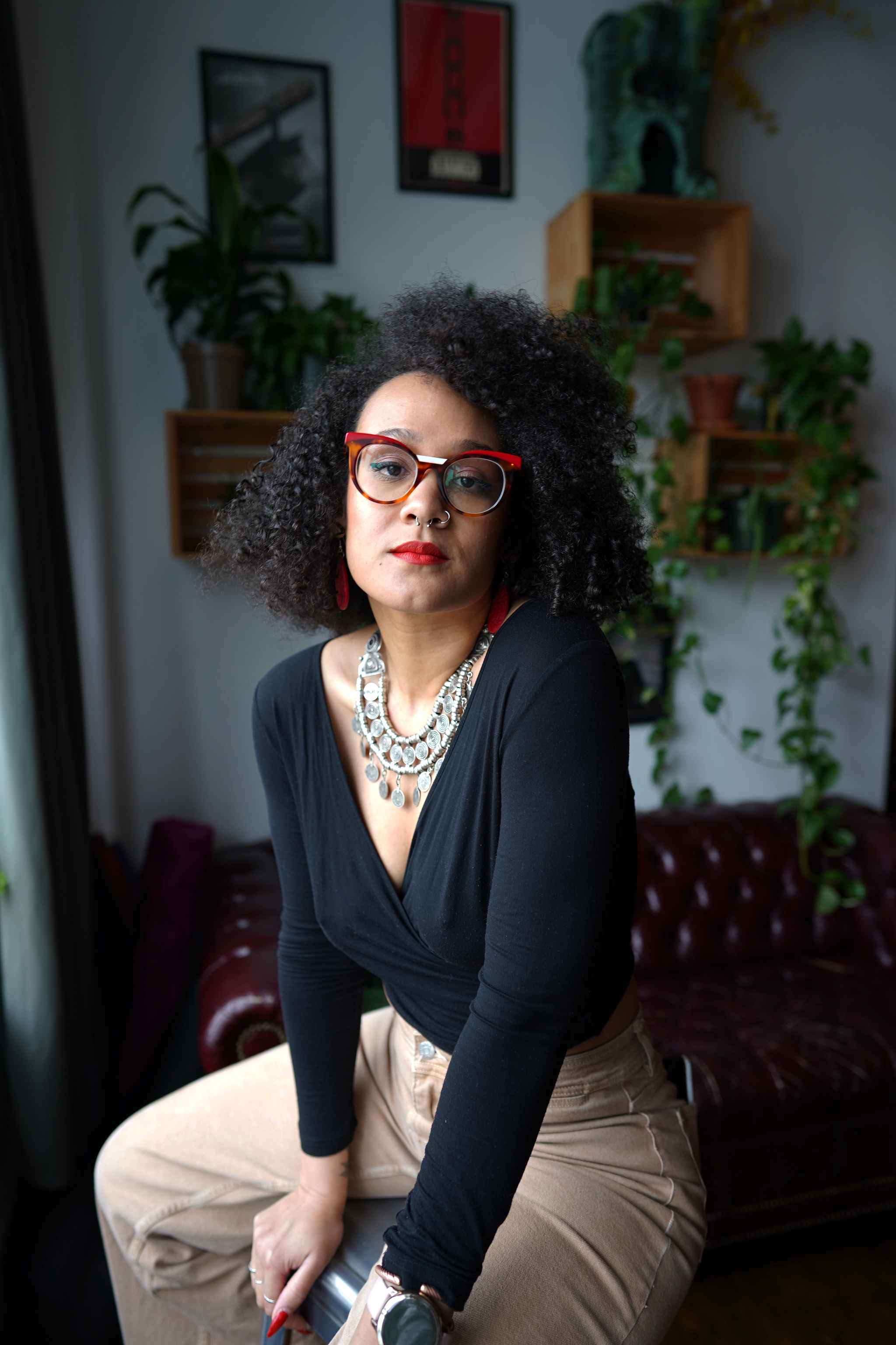 A photo of artist Eleanor Kipping, a Black woman wearing tan pants and a deep v-neck shirt, paired with a large necklace and red-rimmed glasses. She sits in what looks like a living room with plants and a plush leather sofa behind her. 