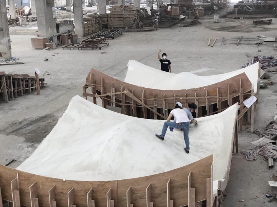 Two students working on a huge structure, with a curved, wooden frame with upright, shelf-like boards along it and a white, undulating surface; a second similar structure is located behind te first. The students are outside on a concrete lot.