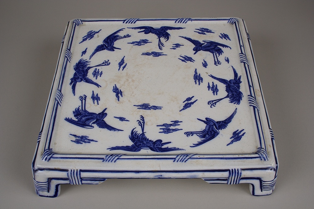 A square bone china base depicting blue storks and clouds forming a circle around the center of the piece.