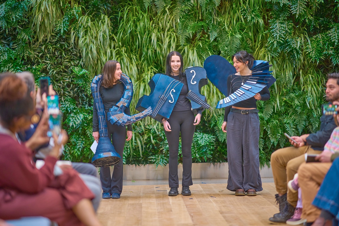 three students stand in front of an audience with a green plant wall backdrop wearing blue music note inspired costumes made from cardboard