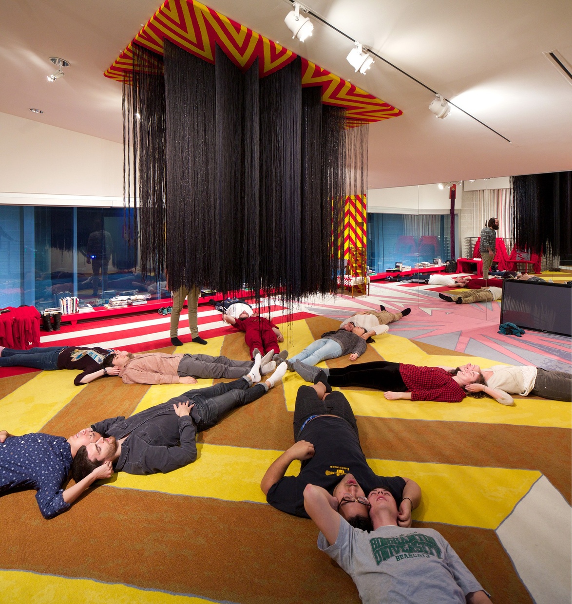 A group of young people lie in a starburst formation on a yellow, abstractly-patterned carpet underneath a red, yellow, and black abstract hanging sculpture.