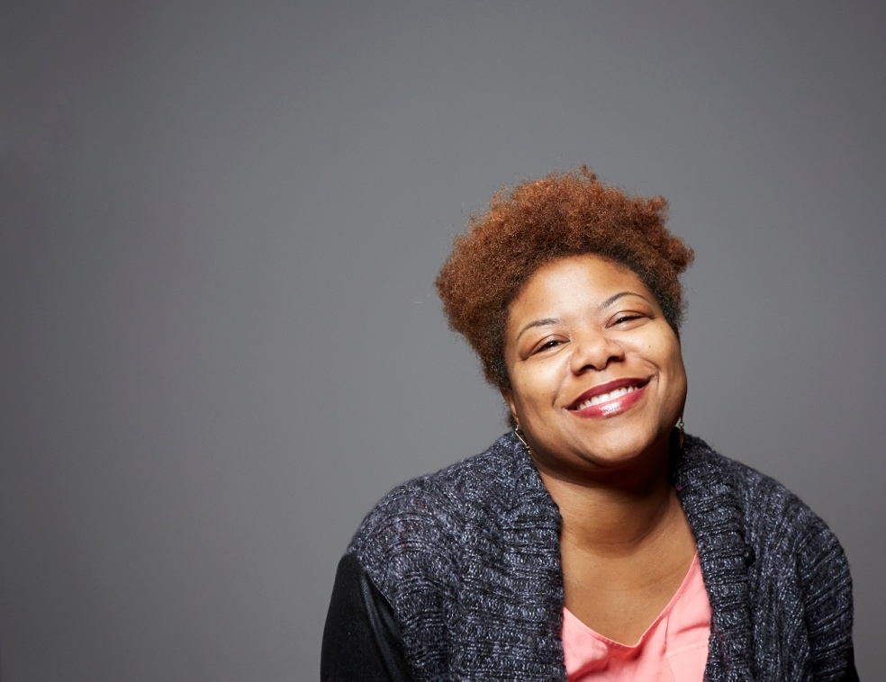 A portrait of Monica L. Williams, a Black woman with short auburn hair. She poses against a gray backdrop and smiles joyfully with her eyes partially closed. 