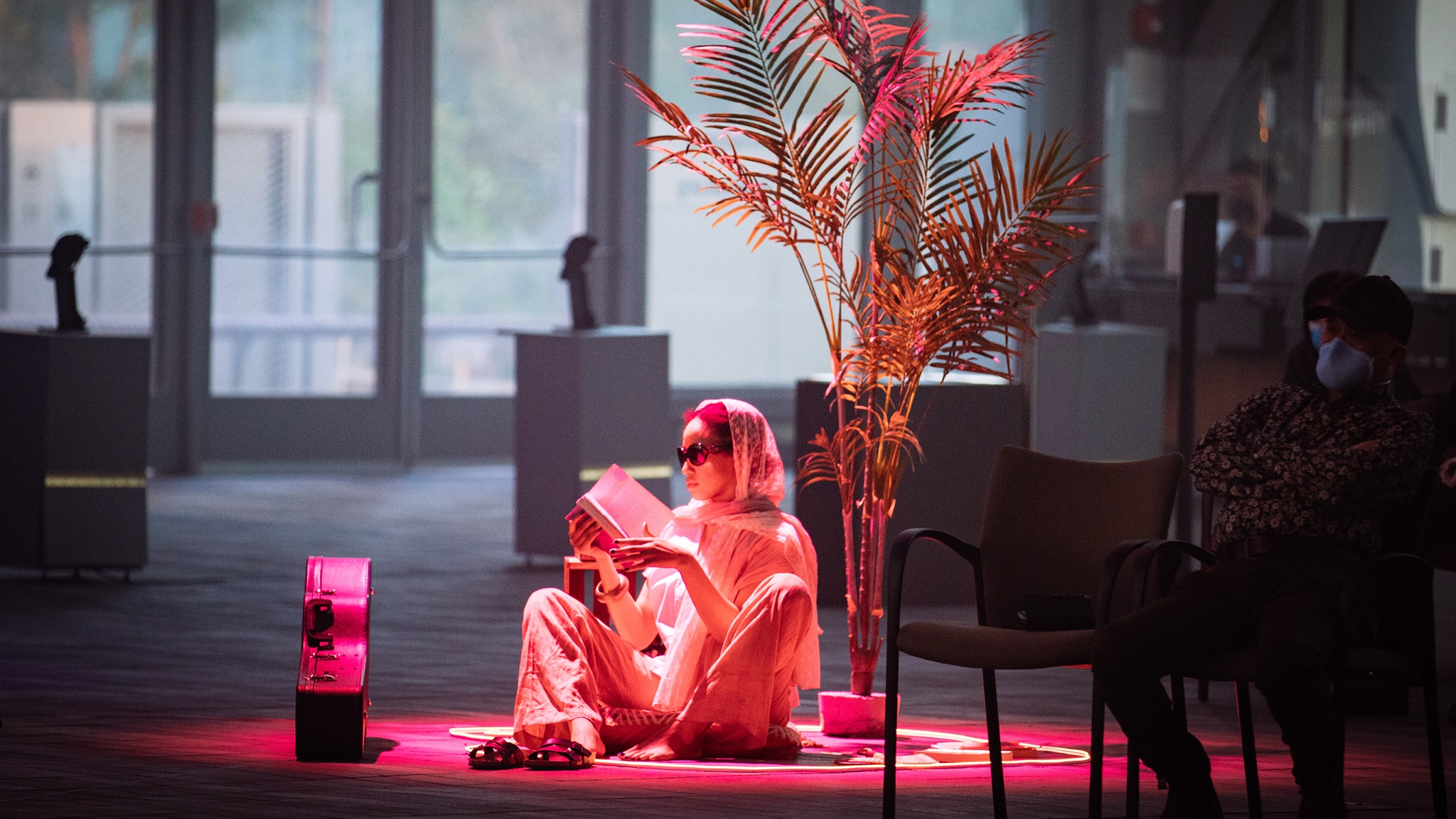 A woman sits on the ground of a performance space illuminated by a red light while she reads a book. Beside her sits a palm-like plant. 