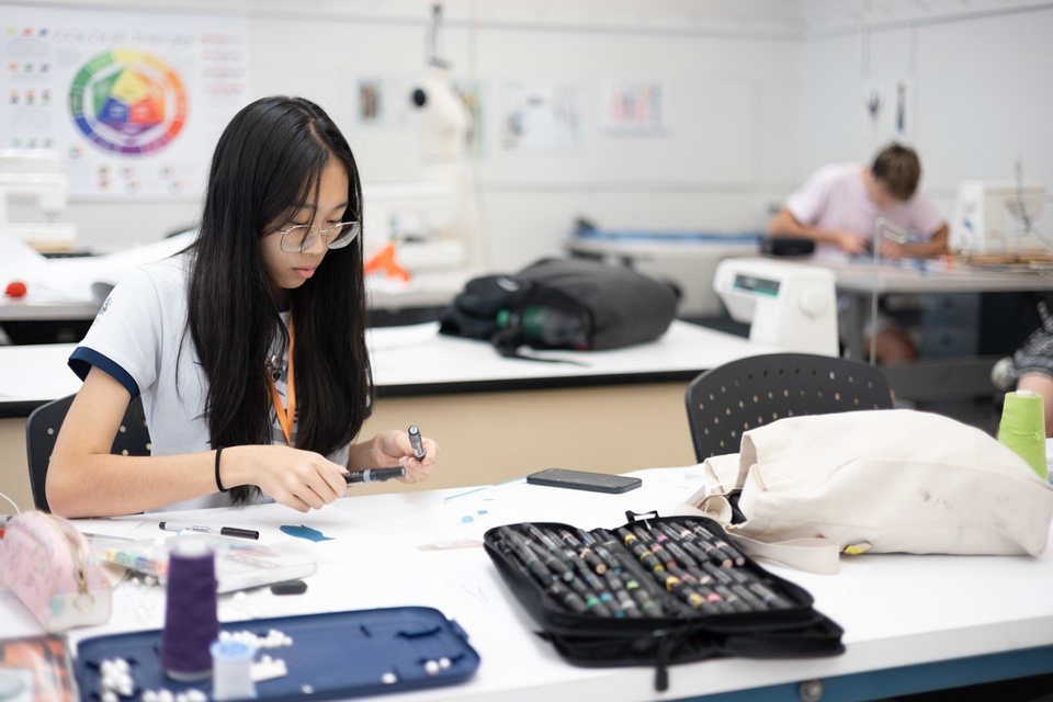 A student with long hair sits at a table working on an art project.