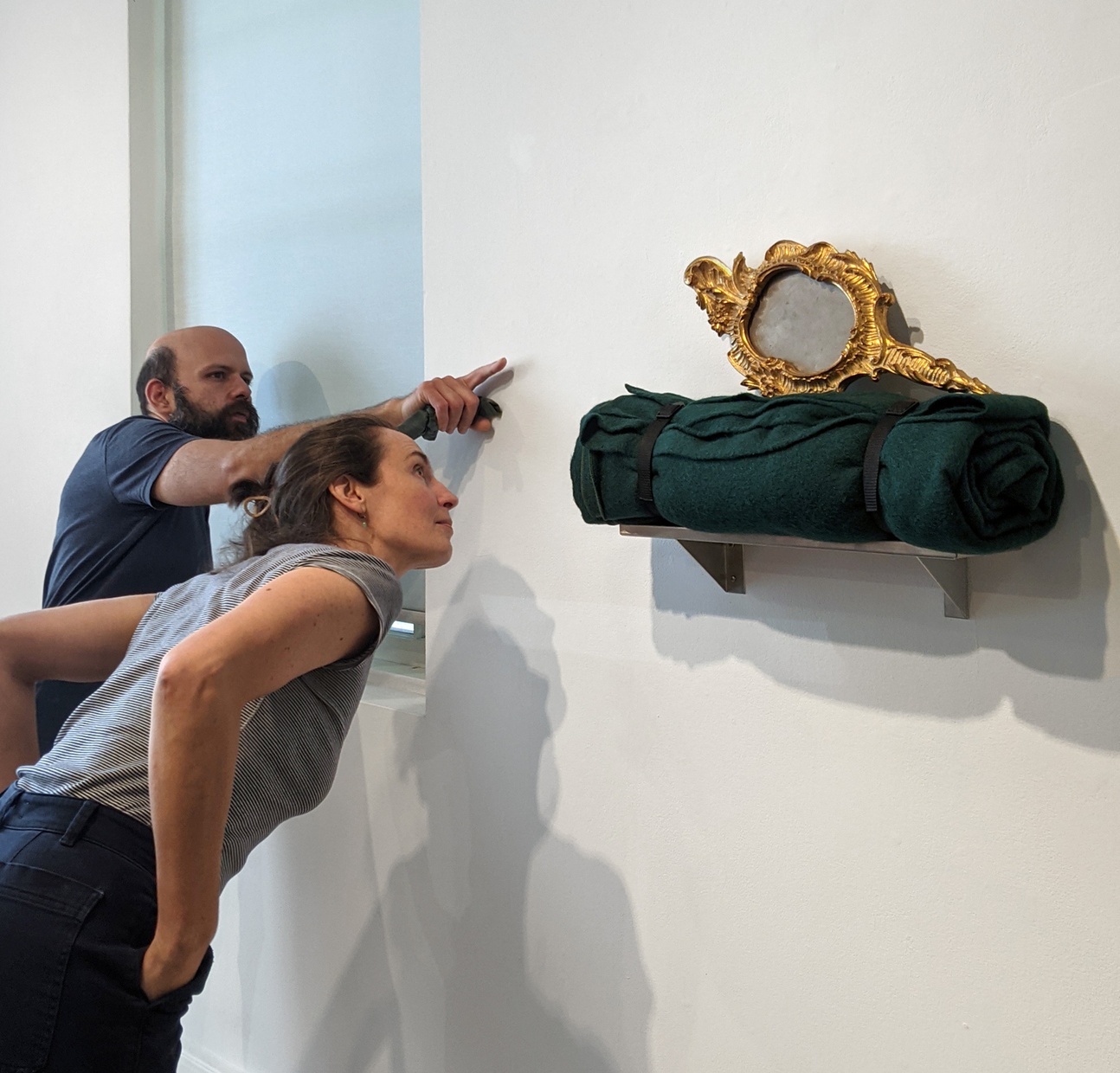 Two individuals, one pointing and the other bending forward, are examining a gold, hand mirror that is laying horizontally atop of a green, rolled-up, blanket.