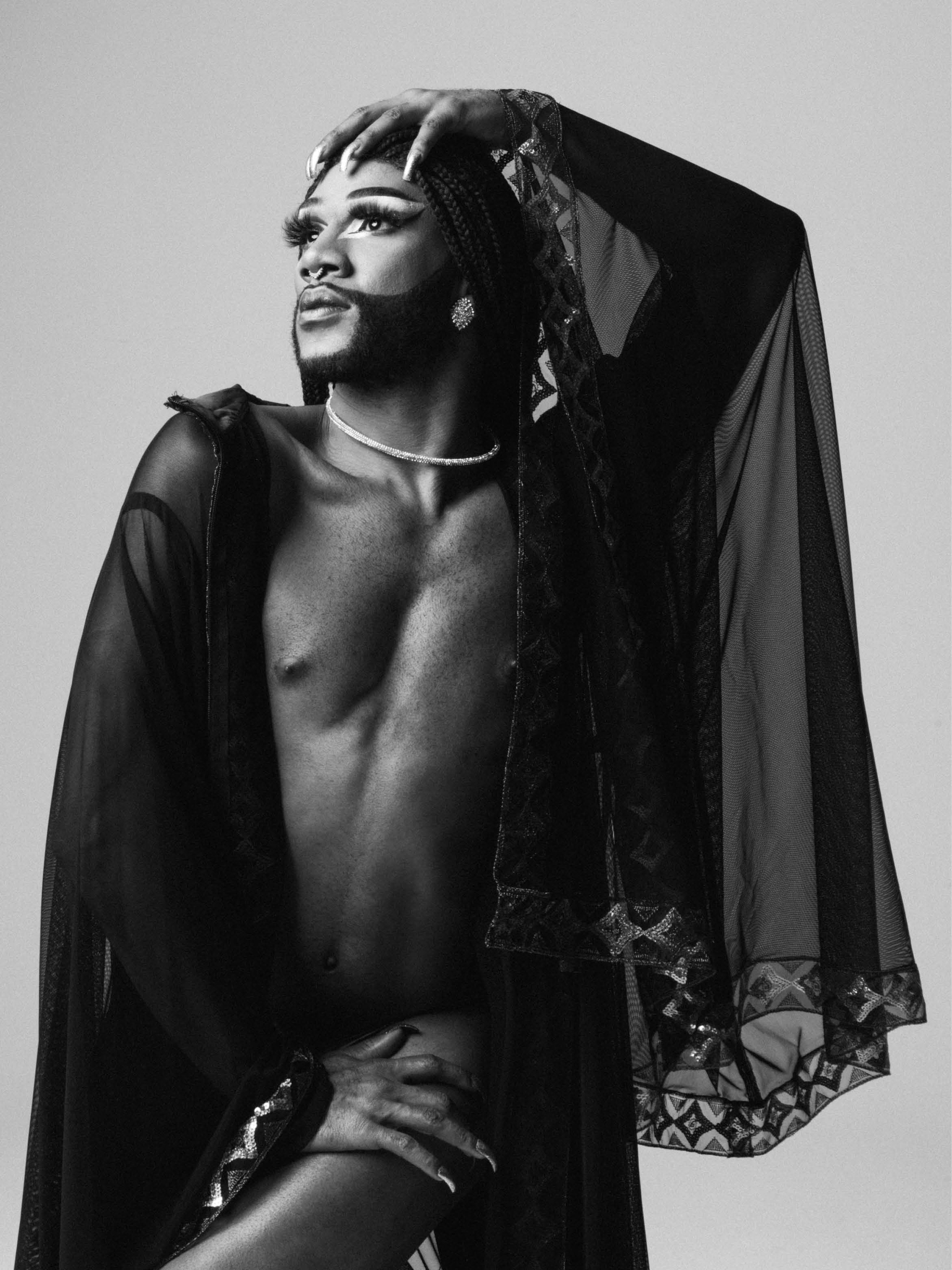 Cain Coleman, a Black multihyphenate drag performer, poses with their body slightly twisted looking dramatically upward toward a light source. They have one hand reached up and resting on the crown of their head and the other reaching down to their left hip. Cain wears a long, sheer black garment that includes a covering worn over the crown of their head, embellished at the edges with gold patterning. Cain has a beard that is styled in sharp contrast to the rest of the smooth skin of their cheeks and face. 