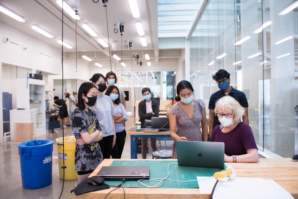 Group of people in a maker space gather around an instructor for a demonstration on a laptop.