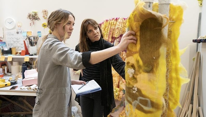 On the left, a woman in gray coveralls stands next to a woman in a black cardigan and black jeans, discussing a large sculpture-in-progress, at right. Each of them stands with an arm extended at shoulder levelto touch the sculpture, which is composed of two 3-inch-wide verticle PVC pipes wrapped in chicken wire and a panel of handmade yellow and brown felt.