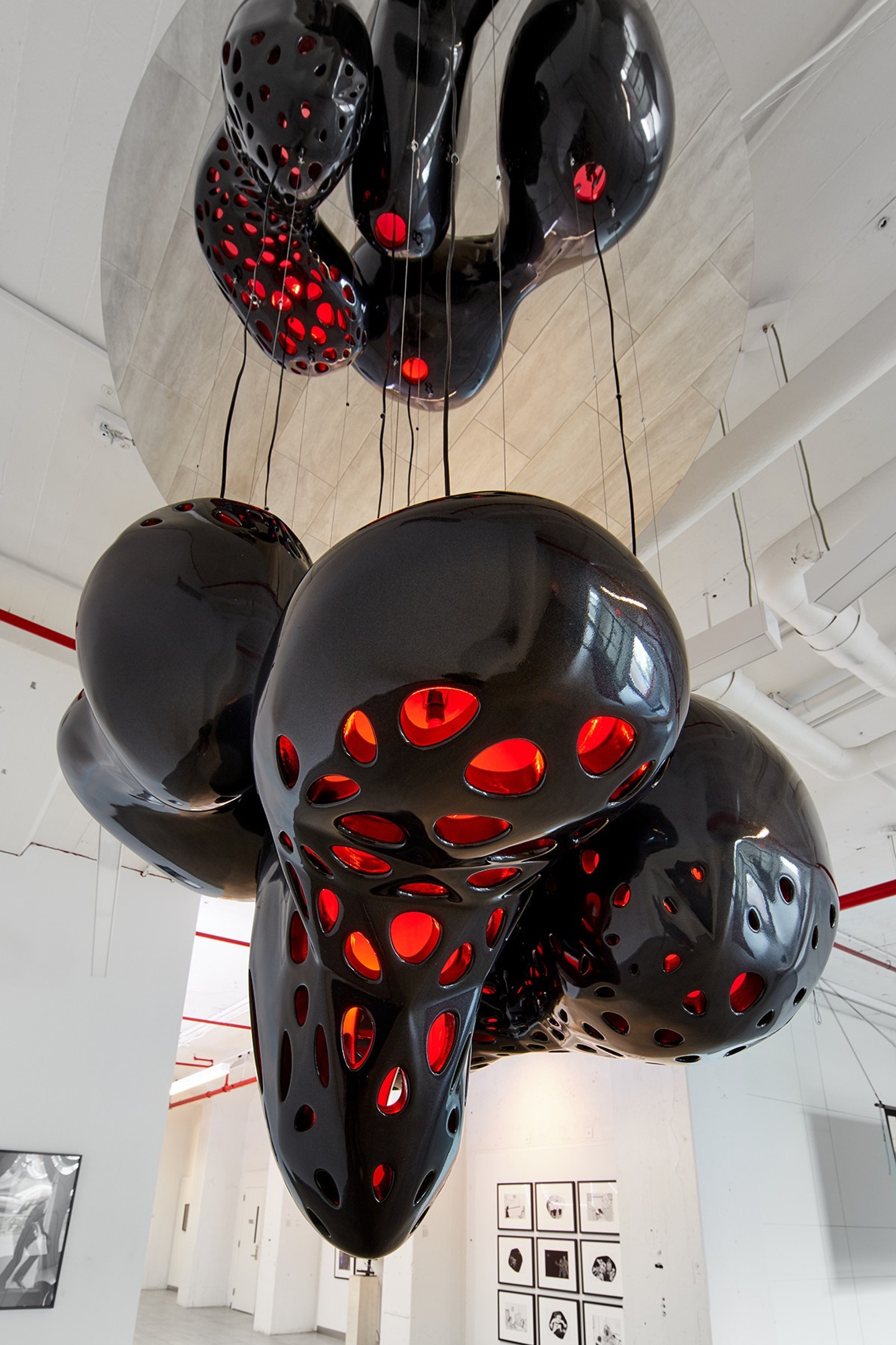 Installation in a white gallery space of two large, bulbous hanging forms connected by black wires/cables/ropes, red light shining through its perforations.