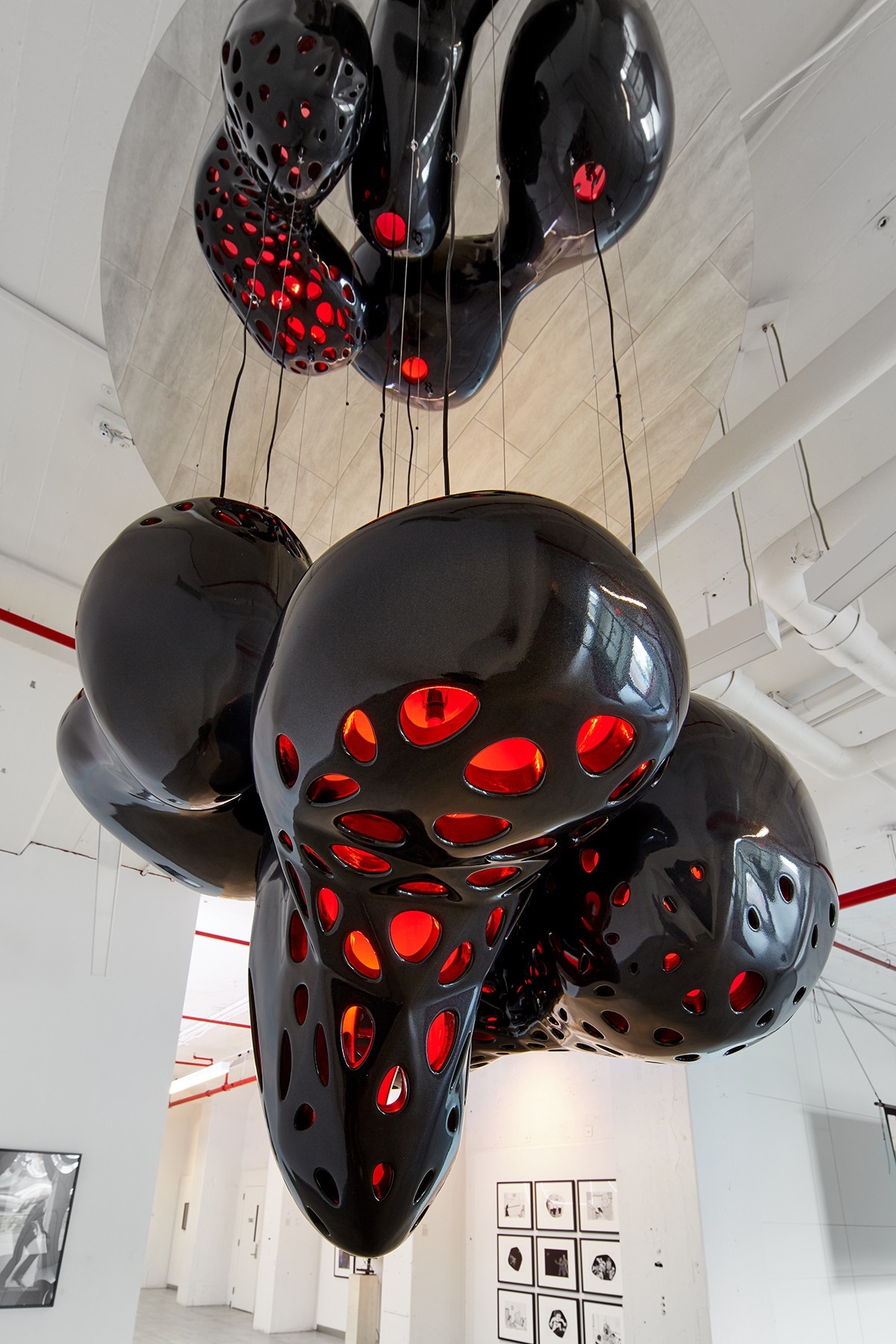 Installation in a white gallery space of two large, bulbous hanging forms connected by black wires/cables/ropes, red light shining through its perforations.