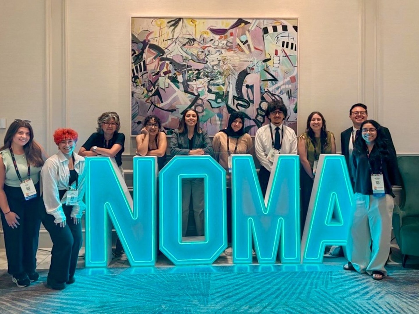 Ten people stand behind an oversized lit blue sign with the letters NOMA.