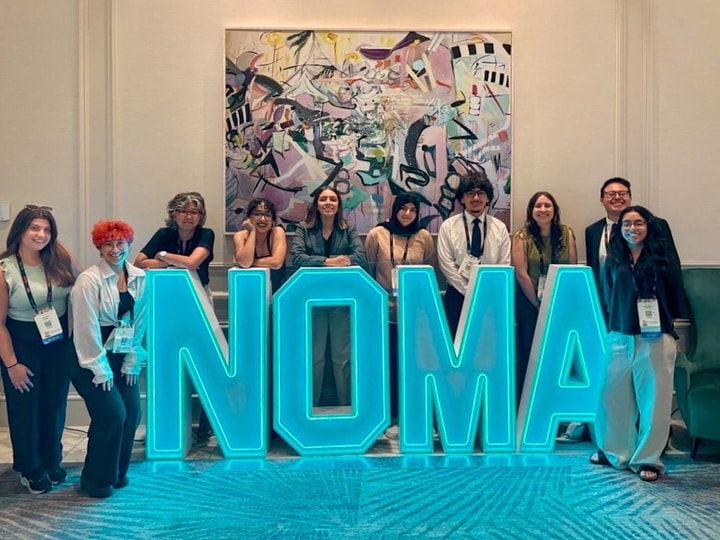 Ten people stand behind an oversized lit blue sign with the letters NOMA.