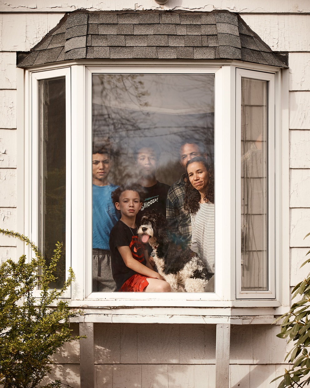 A photograph of a dark-skinned family, an adult male, adult female, and three boys, with a black and white dog looking out a white bay window.