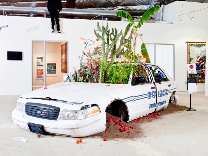 A broken police car sits in the middle of a room. Plants grow out of its windshield, and tomatoes and sand spill out of the wheel wells.  Jordan Weber, “American Dreamers Phase 2,” 2014-15. Installation view. (Photo courtesy of the artist)