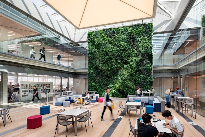 Light-filled interior atrium space with multiple tables and blue and pink soft seating, with people working in the space. The back wall features a floor-to-ceiling wall of green plants; the sides of the two-level space feature floor-to-ceiling glass, looking out into surrounding studio spaces.