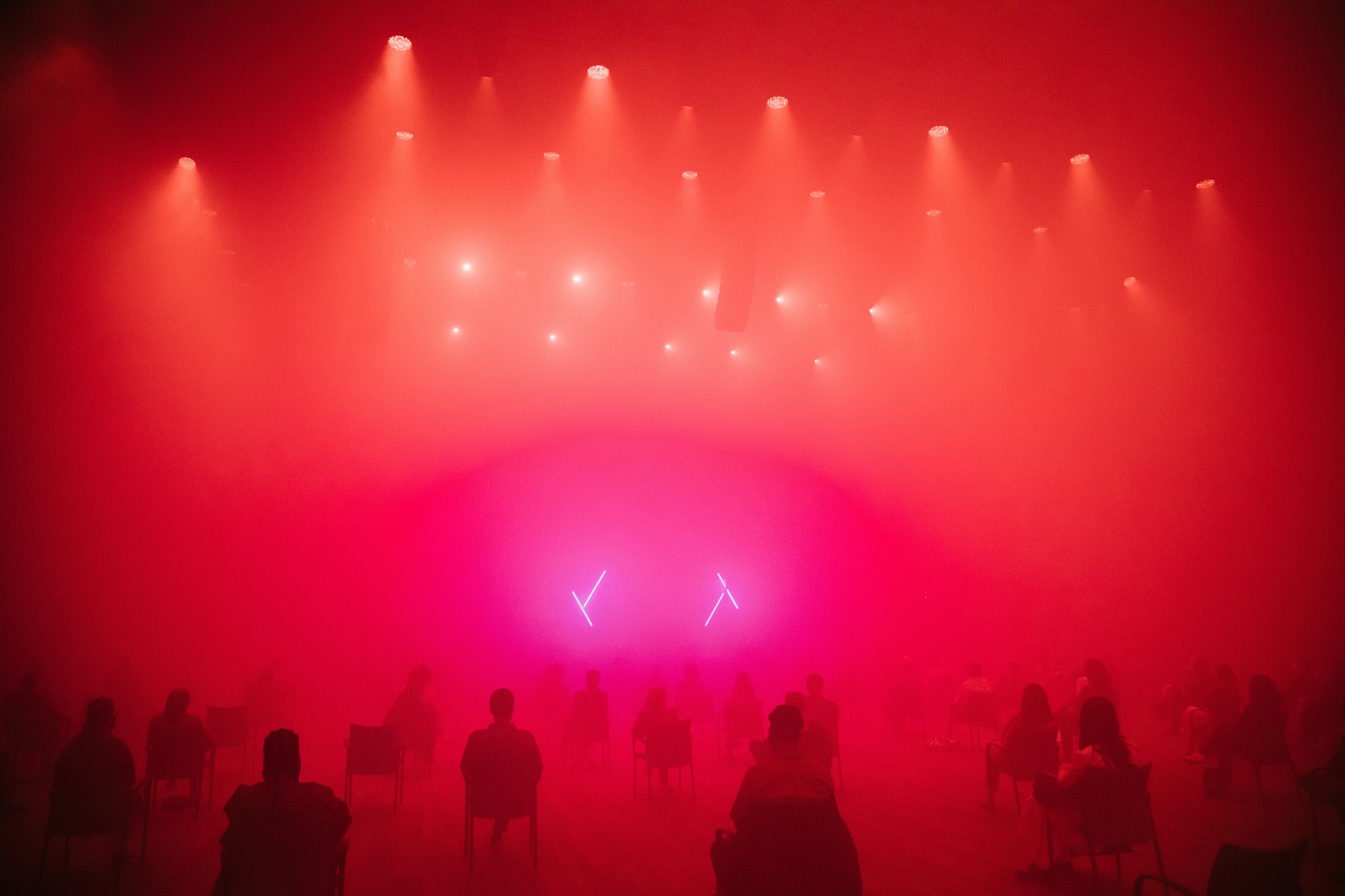 People seated in chairs at a distance from each other across a vast performance space. The space is foggy and atmospheric, lit by orange spotlights and pink fluorescent lights that form two Xs above a stage. 