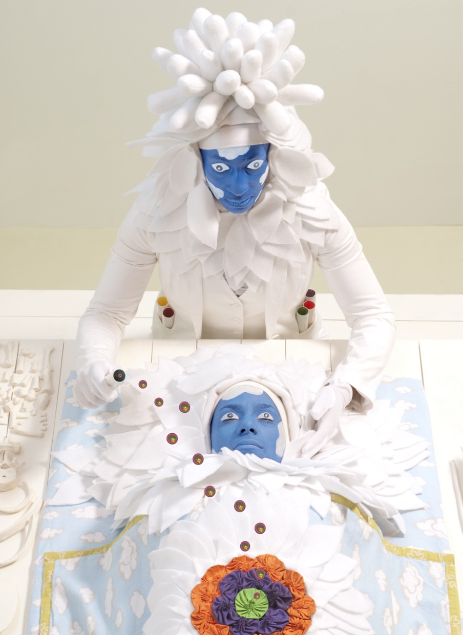 A person with a painted blue face in a white, plant-like costume looks down on a similar person lying under a blue blanket with clouds and a flower embroidered on it.