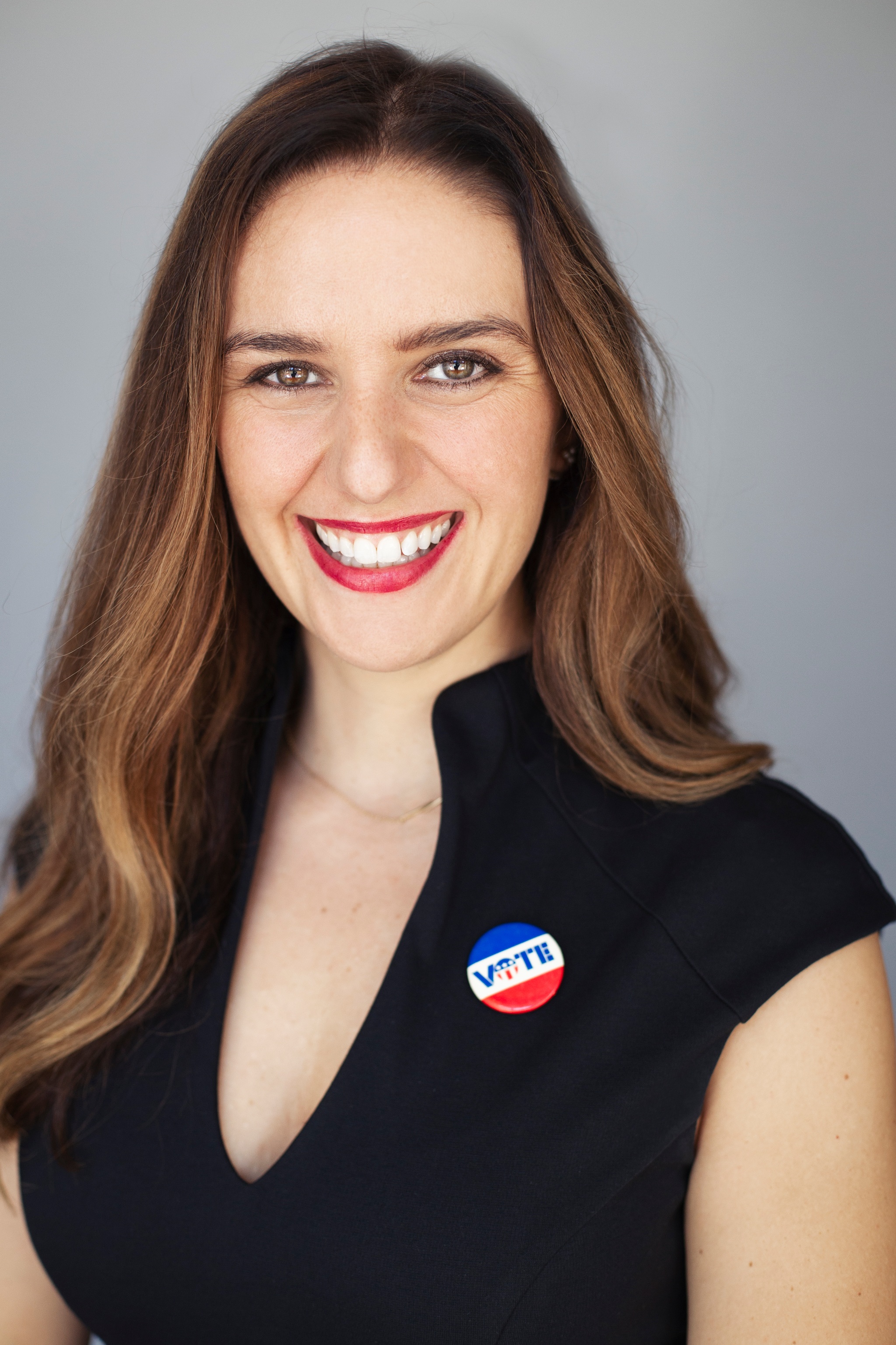 Molly McGrath smiles at the camera. She has brown hair that falls on either side of her head and wears a black blouse with a “Vote” button in red, white, and blue just below her left shoulder. 
