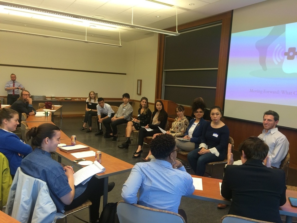 A group of people sit in a row facing another group of people. A presentation screen appears behind them.