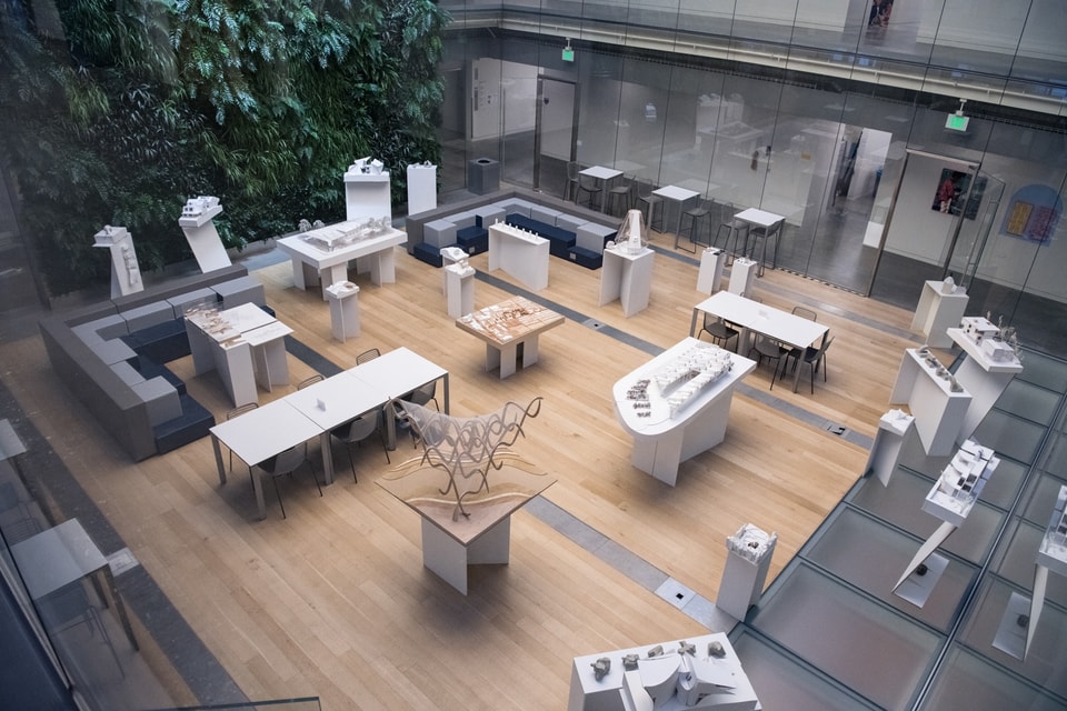 Overhead view of two dozen architectural models on white stands arranged around an interior courtyard with a living green wall on one end.