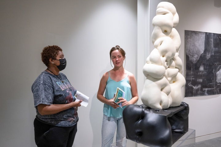 Emily Elhoffer speaks with Allena Brazier in front of her fleshy white and black sculpture on top a transparent pedestal  