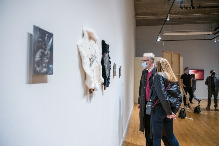 Two people look at a gallery wall on which is hung a steel plate and two rabbit pelts in black and white. The plate has an engraving of a rabbit and the two pelts contain drawings that are not legible.