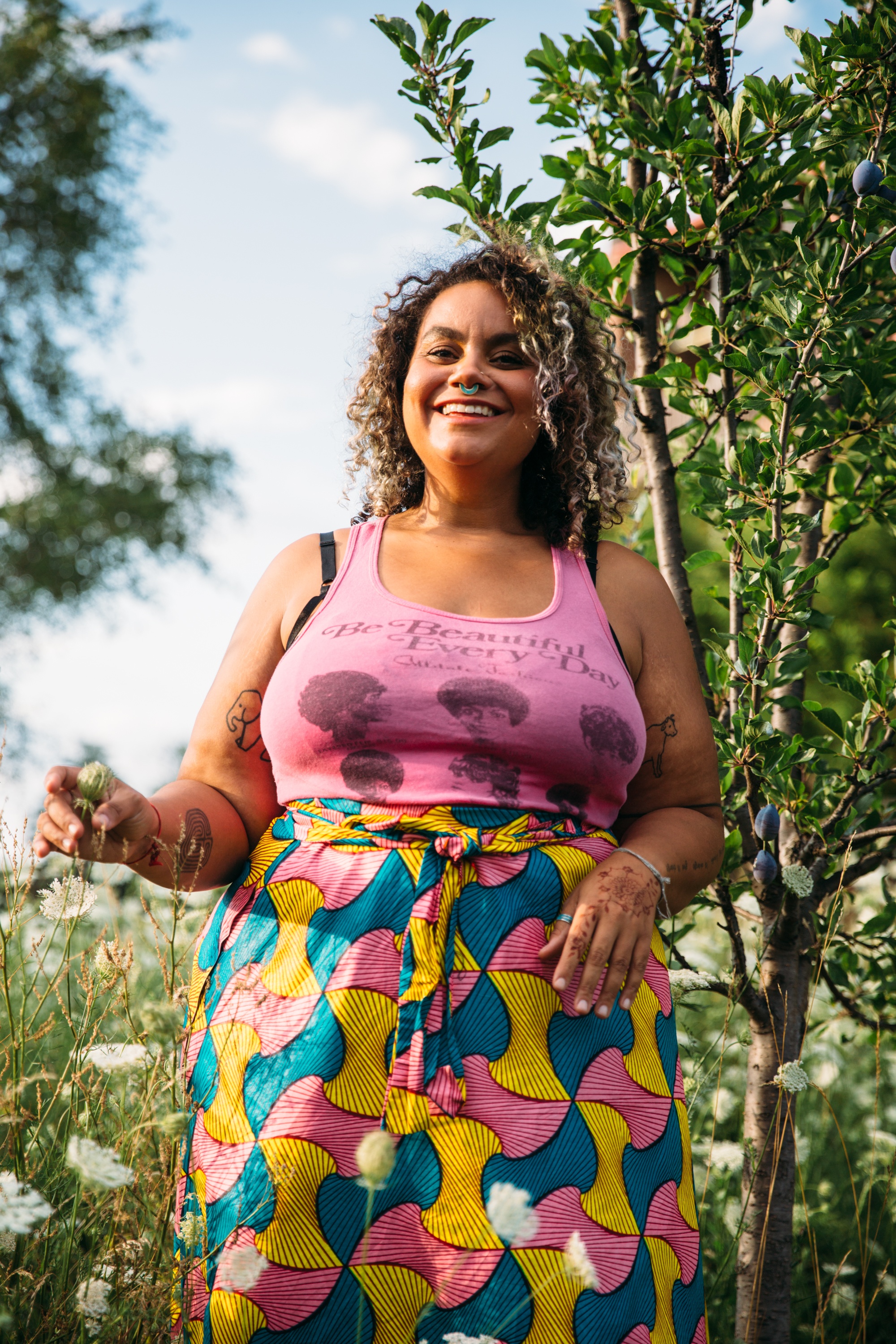 A portrait of adrienne maree brown standing outside surrounded by grasses, flowers, and small trees. She smiles broadly and wears a pink tank top and colorful patterned skirt. 